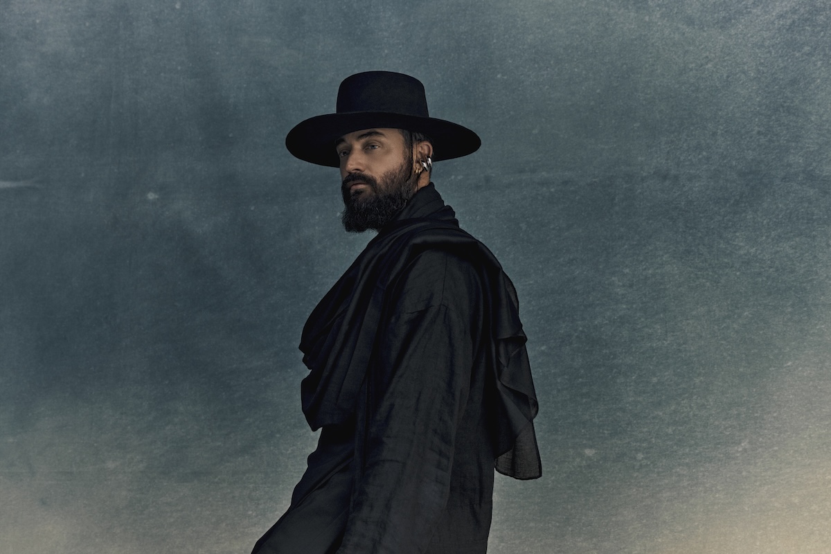 Musician RY X posing in profile against a textured blue backdrop, wearing a wide-brimmed black hat, dark draped clothing, and a full beard.